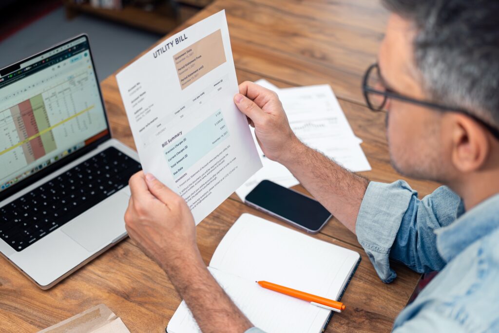 Grey haired tenant man holds printed utility bill at table with notebook and laptop debts growth